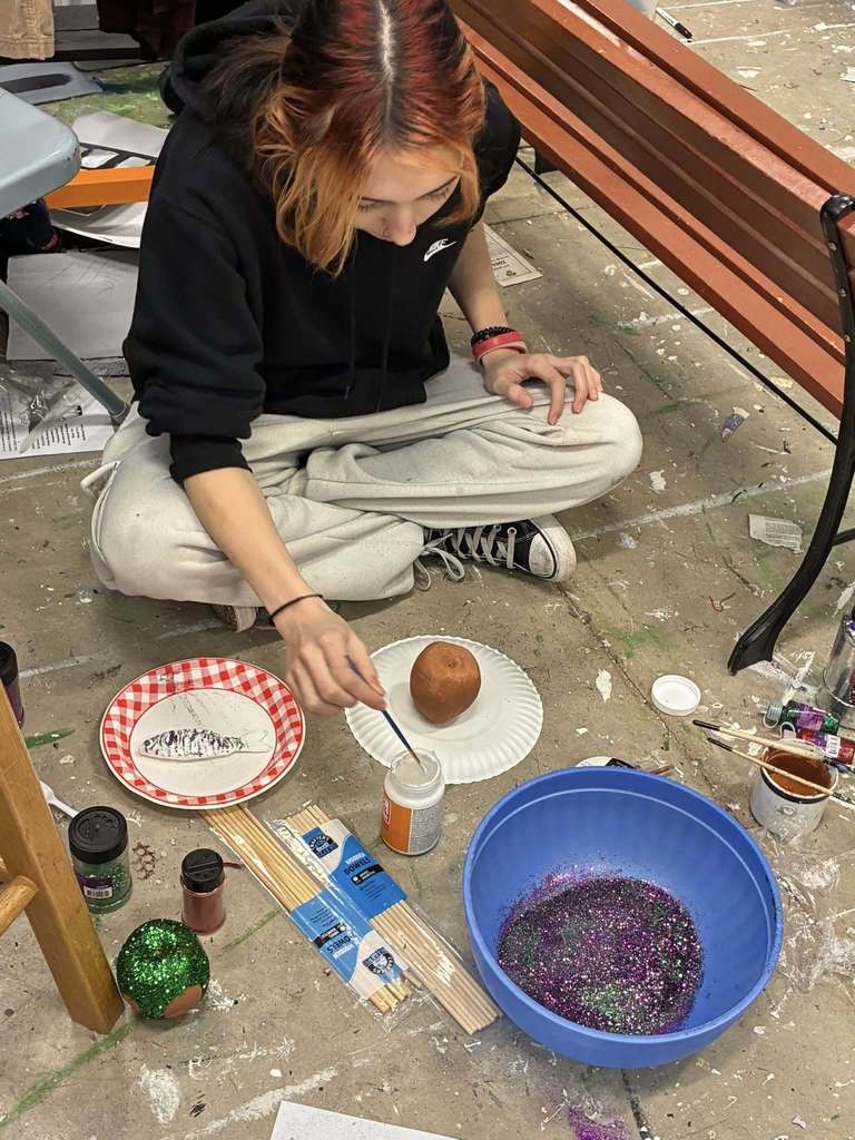 Person kneeling, painting a potato on a plate with various paint supplies and a blue bowl nearby.