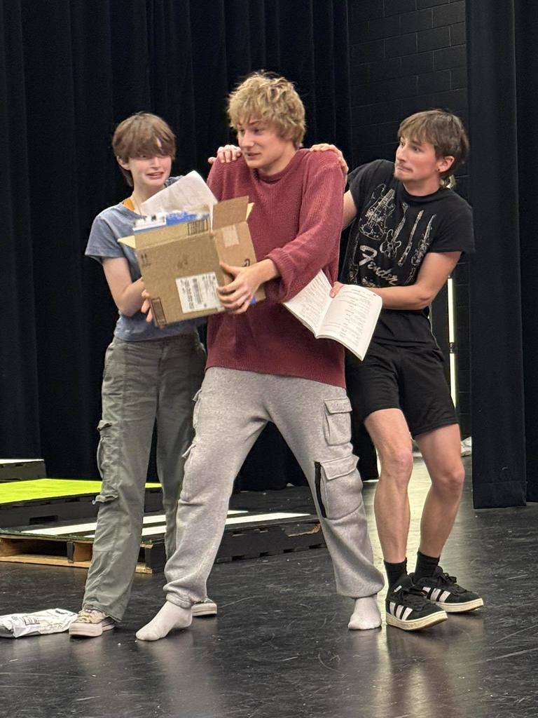 Three young individuals stand on a stage, one holding a cardboard box, another with a book, and the third supporting the one with the box.