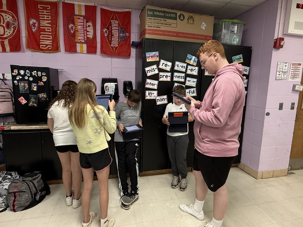 Five students in a classroom using tablets, one student is standing with tablet. Purple walls and banners are in background.