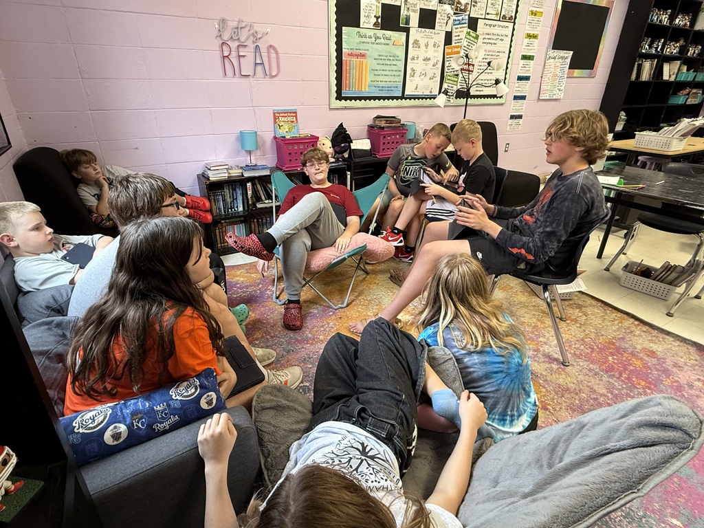 Kids sit in a circle on floor cushions and chairs in a room with pink walls and bulletin boards.