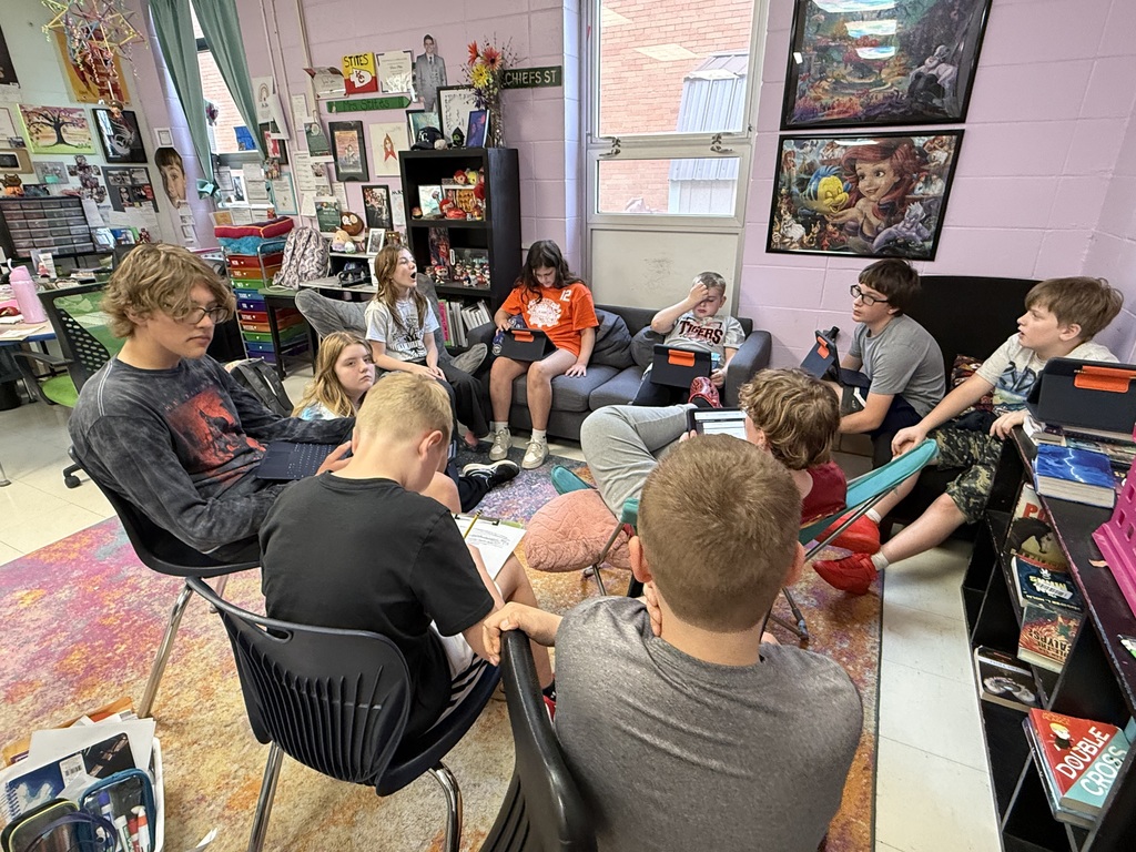 People in chairs around a room. Some are sitting on a couch. A bookshelf is nearby.