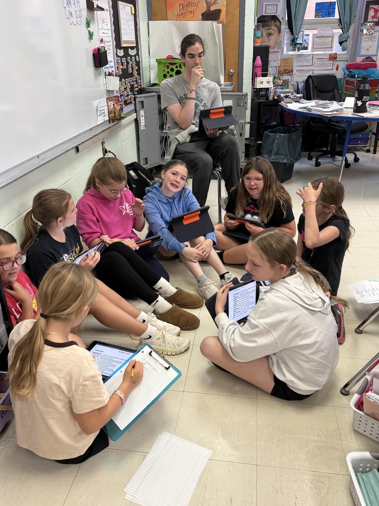 Students sit in a circle on a floor, using tablets and papers, with a teacher seated in the background.