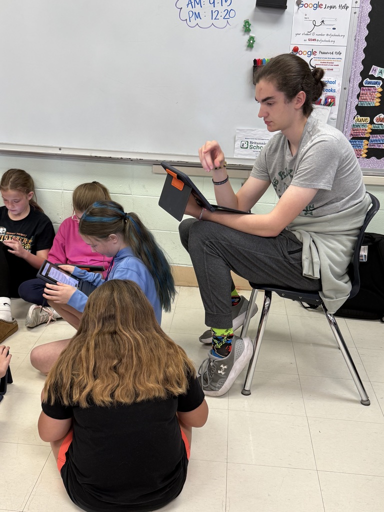 A woman sits on a chair with students around her; they all hold tablets and look at the screens.