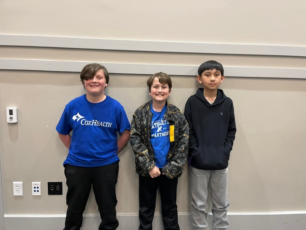 Three boys stand together in front of a wall. One wears a blue shirt, another a camouflage jacket, and the last a dark-colored hoodie.