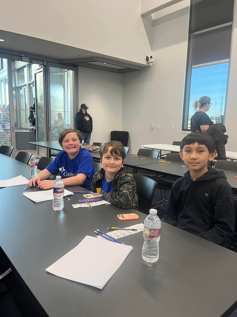 Three children sit at a long table with pens, paper, and water bottles in a classroom.