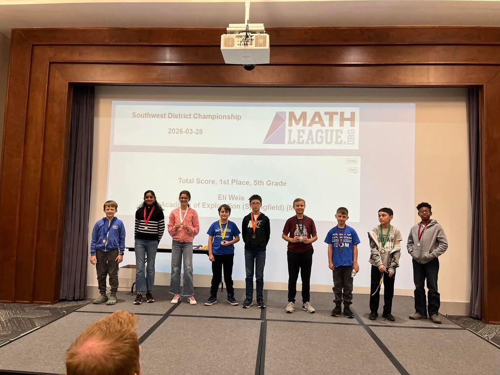 Children stand on a stage holding medals. A projector screen shows Math League text. Wooden walls and curtains frame the stage.