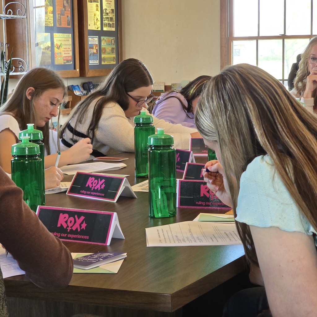 Five people seated around a table, writing on papers with green bottles. A woman in a white shirt with long hair leans forward.