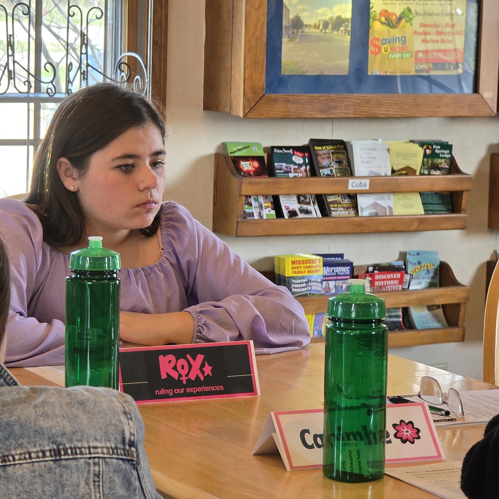 A young woman sits at a table with two green bottles and a sign reading RoX+.