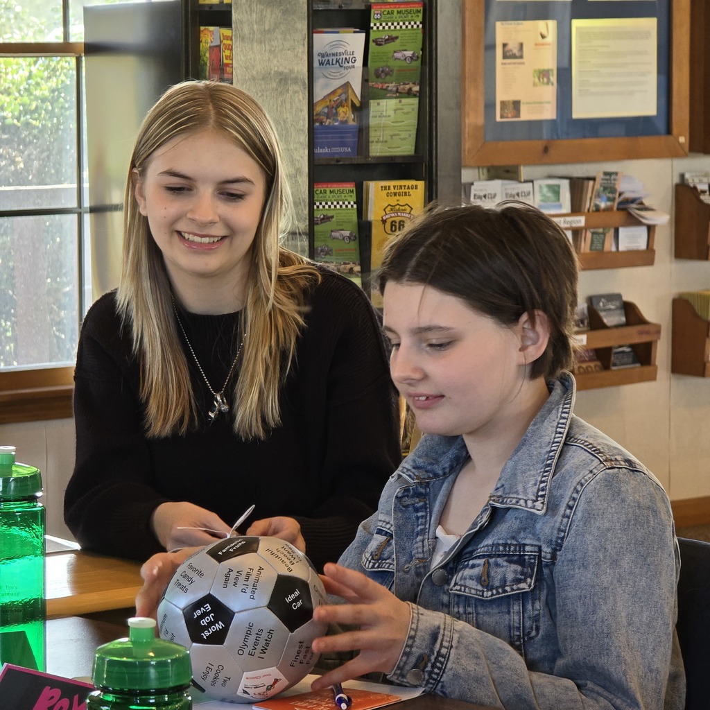 Two women sit at a table; one holds a soccer ball while the other writes on it.