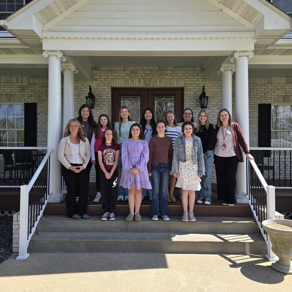 A group of people stand on a porch with steps, a brick wall, and decorative columns.