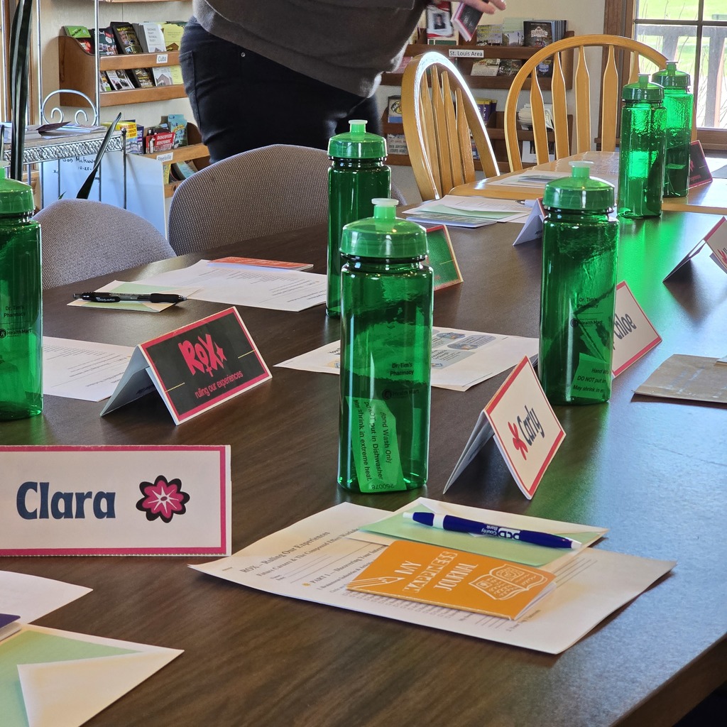 A table with name cards, pens, and green water bottles. A person in gray stands behind the table.