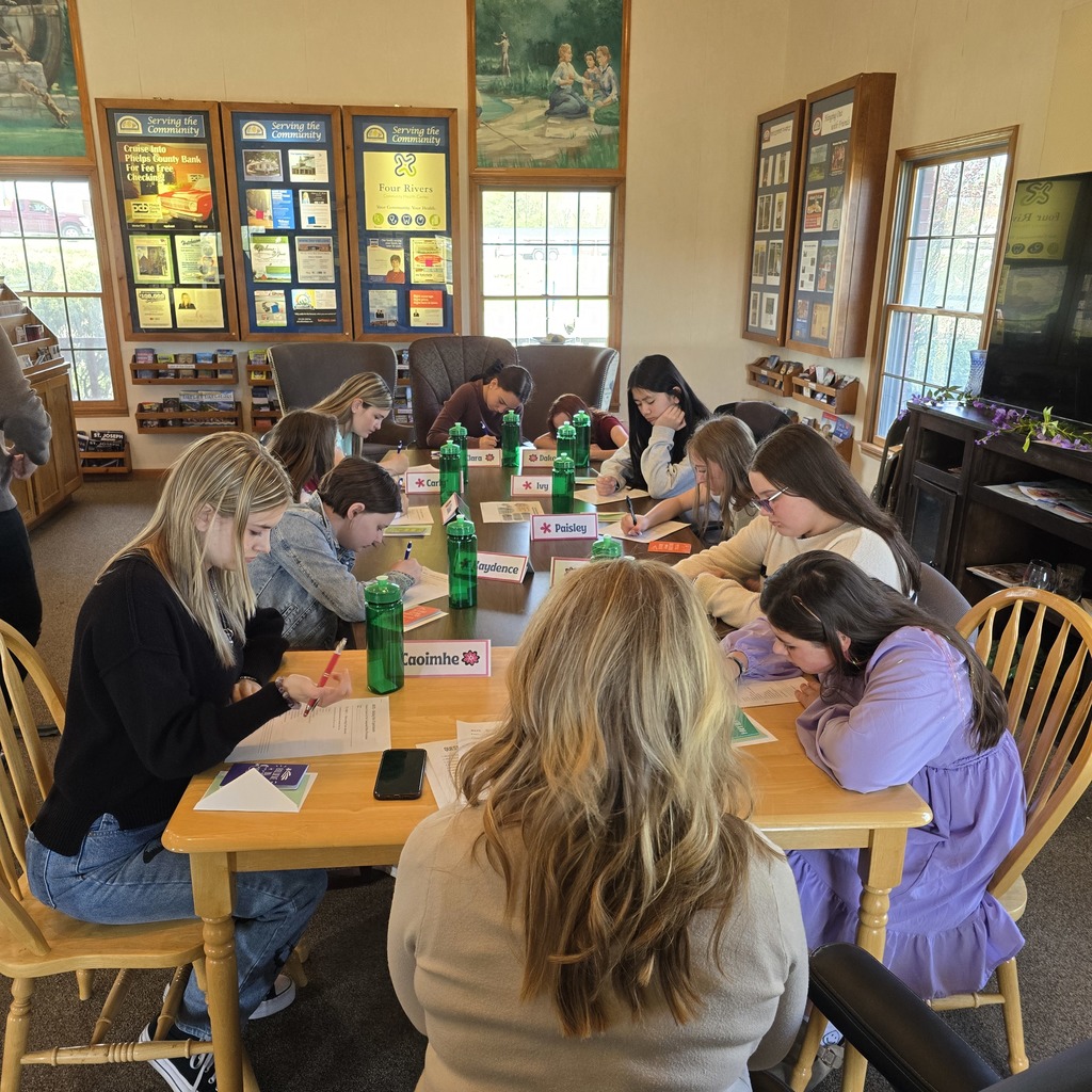 A group of people sitting around a table, writing with pens on paper, in a room with windows and framed pictures on the wall.