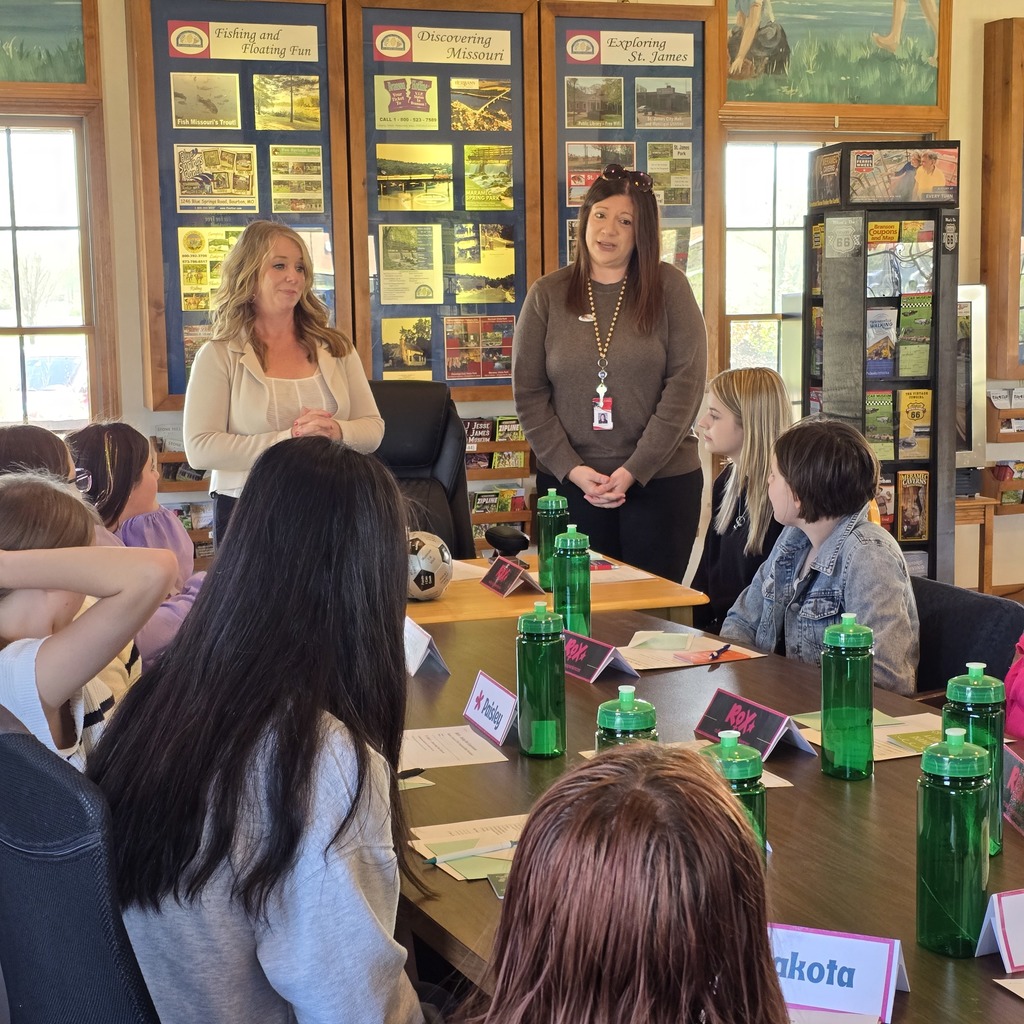 People in a classroom. Two women stand at the front. A table has water bottles.