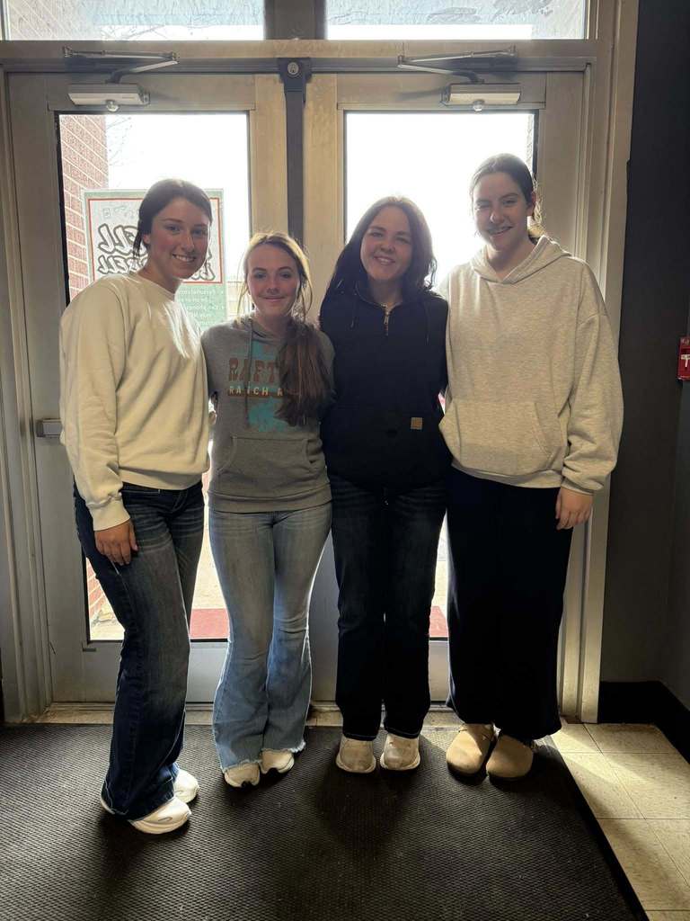 Four individuals stand next to each other in front of a door. Two wear white sweaters, one wears a black jacket, and one wears a beige hoodie.