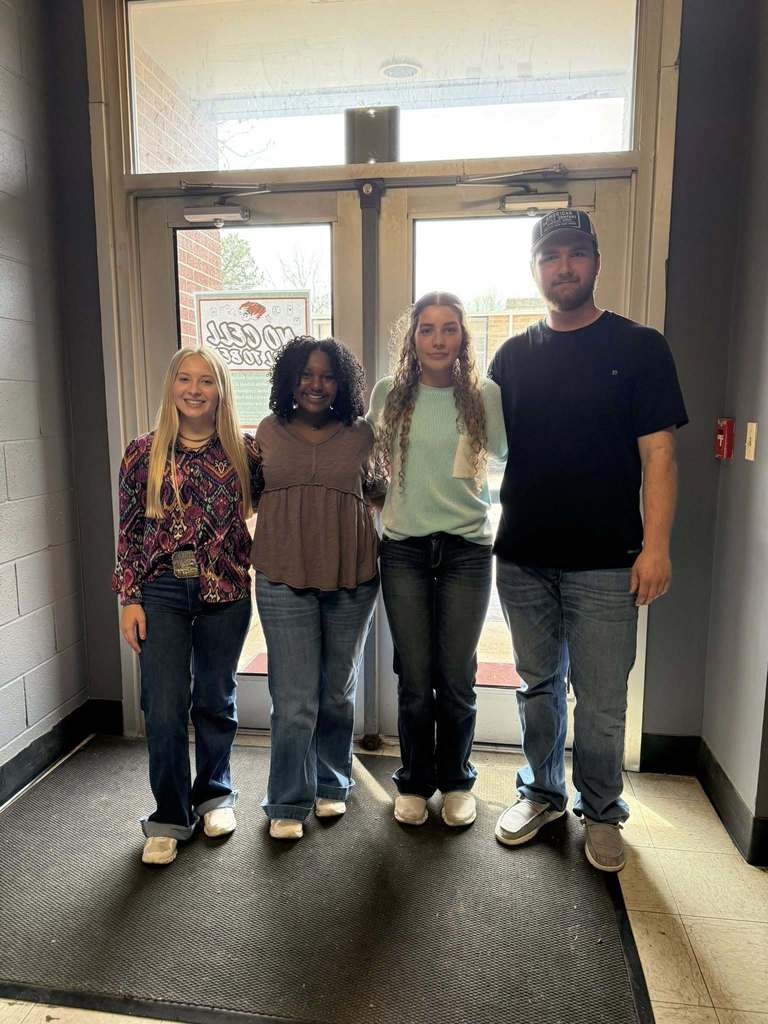 Four individuals stand together in front of a glass door, smiling. Two women wear jeans, one with a blouse, another with a t-shirt. A man wears a cap and a t-shirt.
