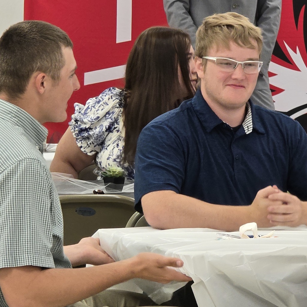 People seated around a table, including two men facing each other, one with glasses. A woman behind them with a floral shirt.