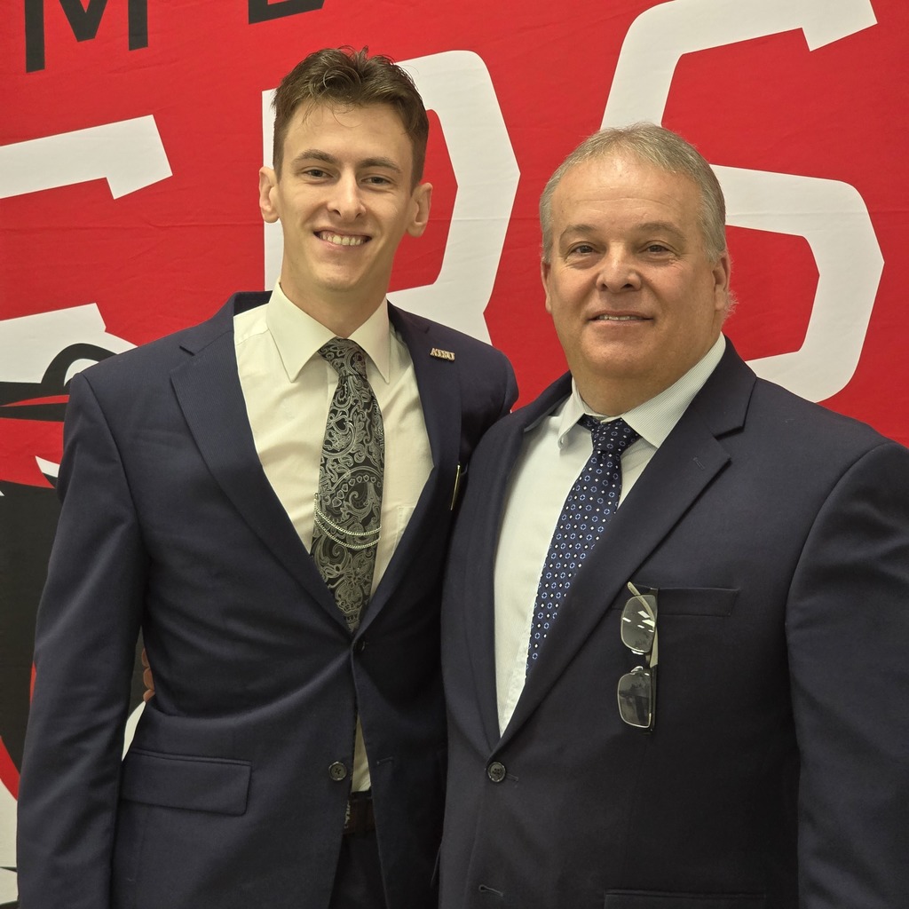 Two men stand next to each other in front of a red backdrop. One wears a patterned tie; the other has glasses on his shirt.