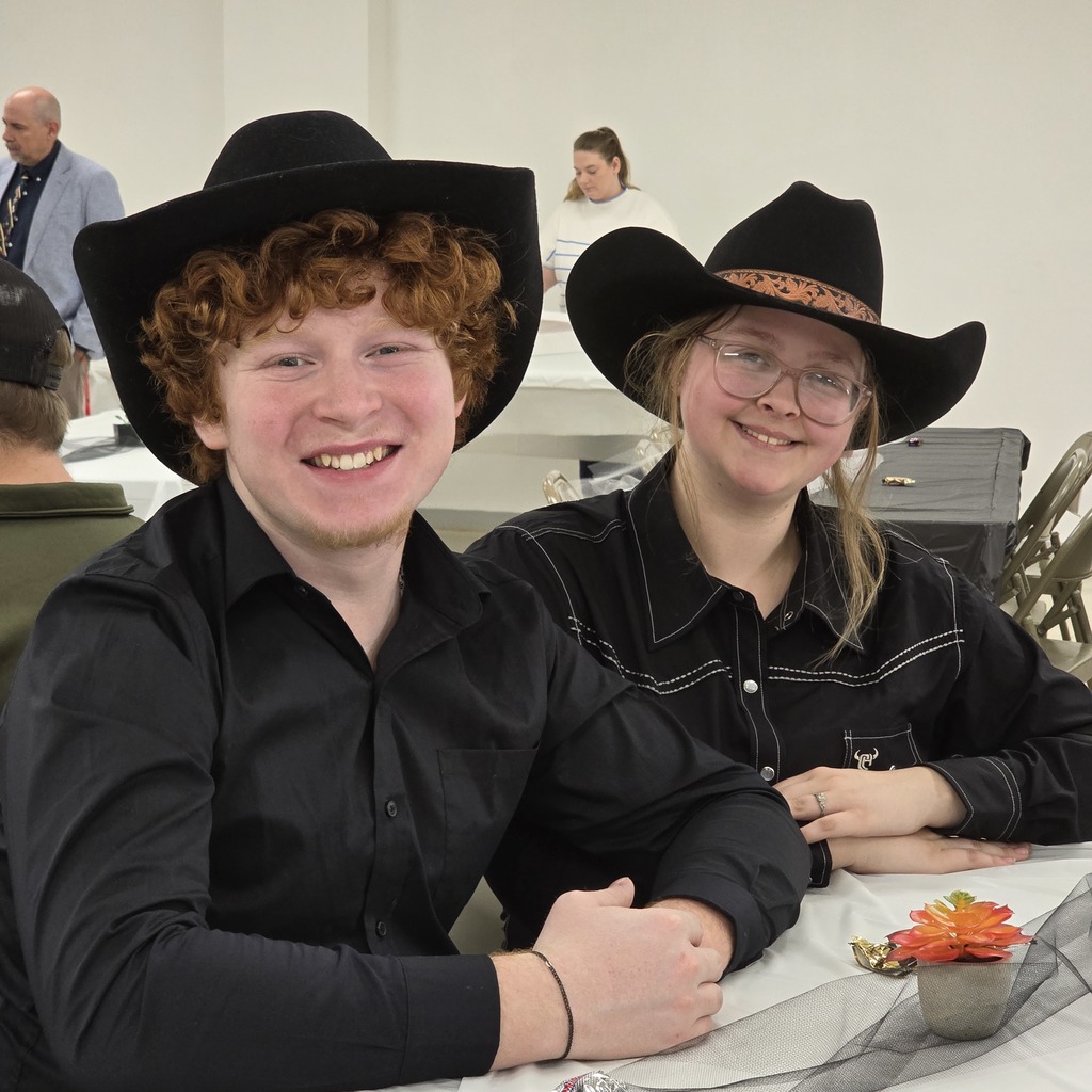 Two individuals wearing black cowboy hats smile at the camera. A small potted plant is on the table.