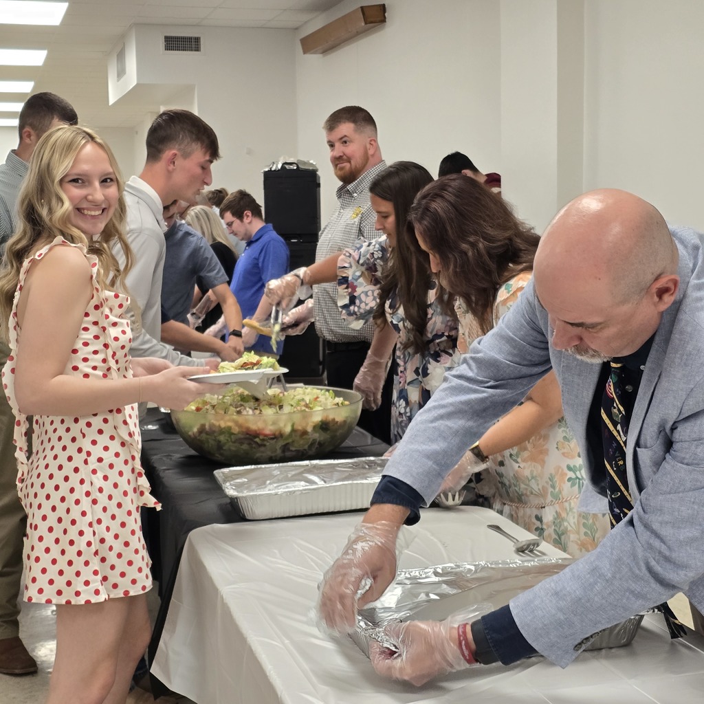 A group of adults in a room gather around tables. One person in a polka-dot dress serves food.