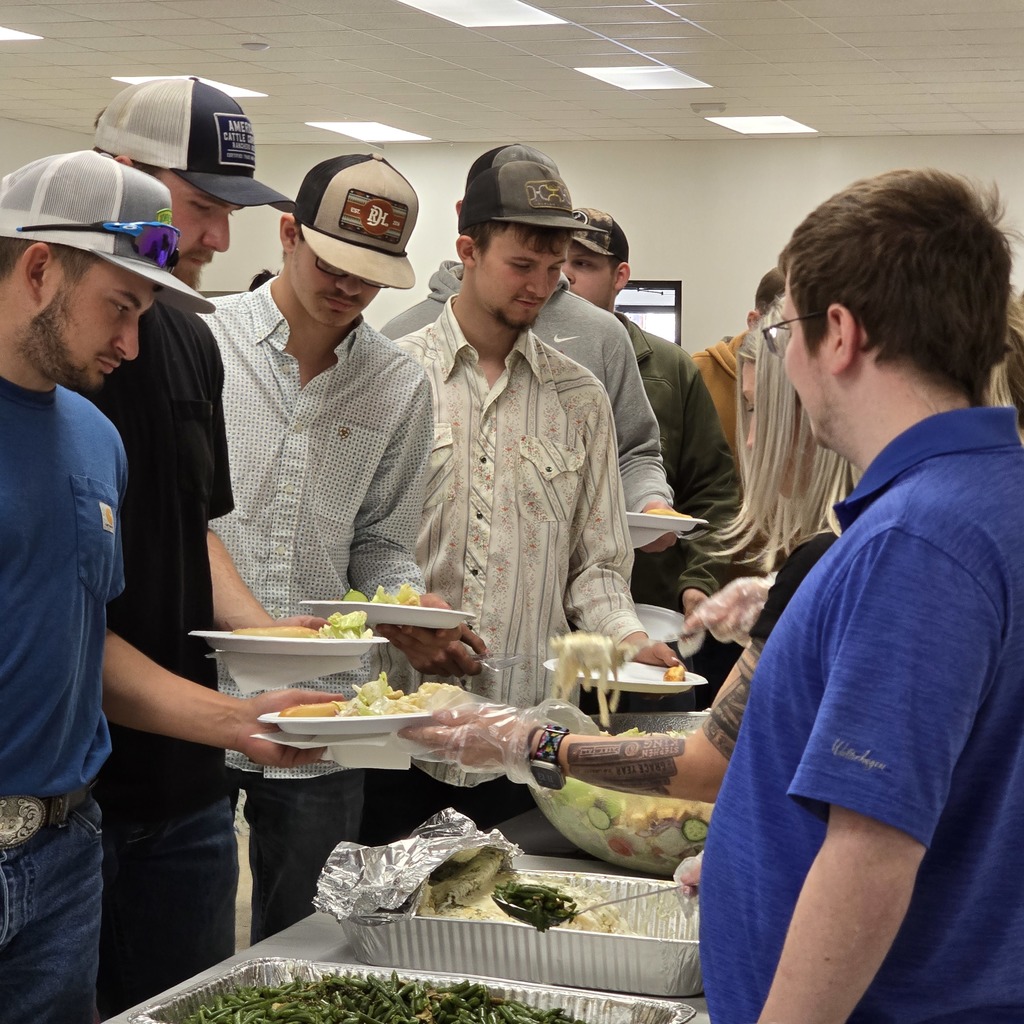 People in a room with light overhead gather around a table for a meal. Trays of green vegetables and a salad are present.
