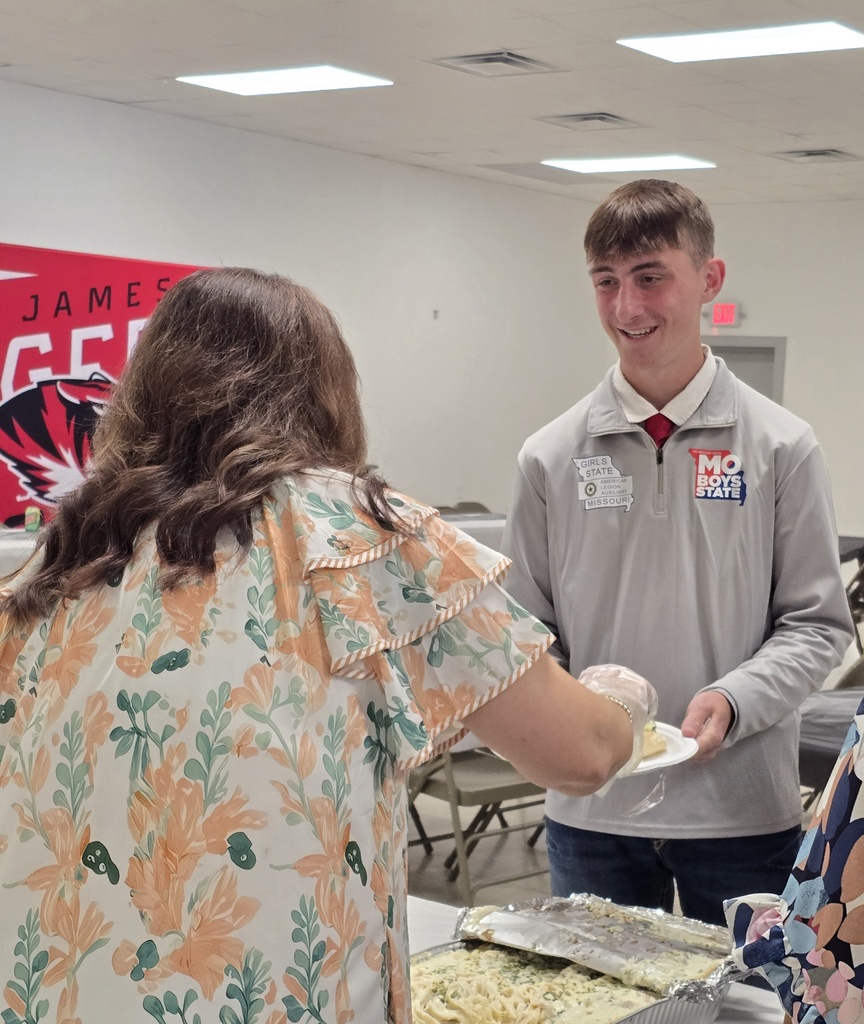 A man and woman serving food at a table. The man is in a gray jacket. Behind them is a red banner.