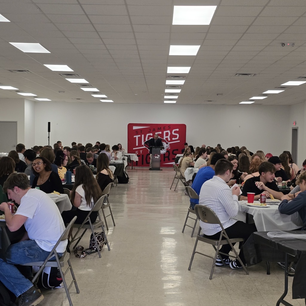 A large room with many people seated at tables. White walls, ceiling lights, and a red sign reading "Tigers" are visible.