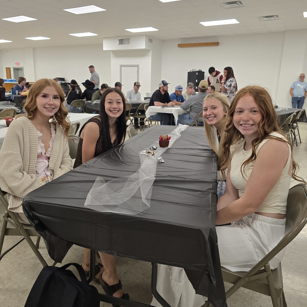 Four women sit at a table in a room with a white ceiling, a black tablecloth, and others dining.