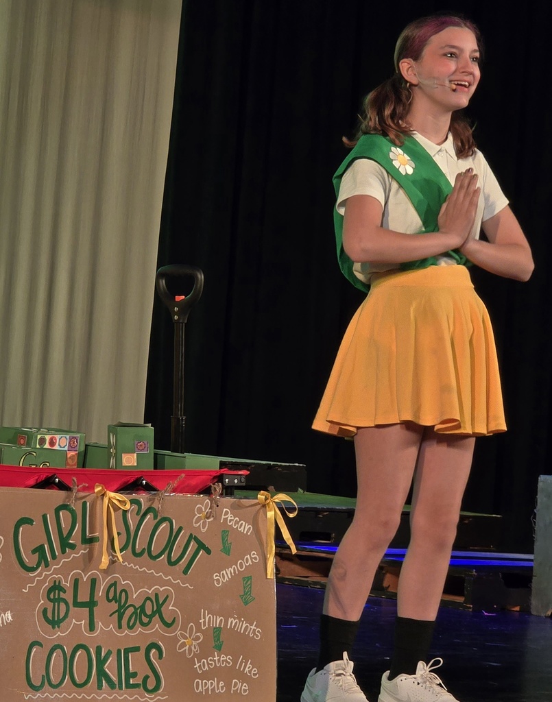 A woman in a yellow skirt and white shirt stands with hands together. Behind her is a sign reading "GIRL SCOUT COOKIES."