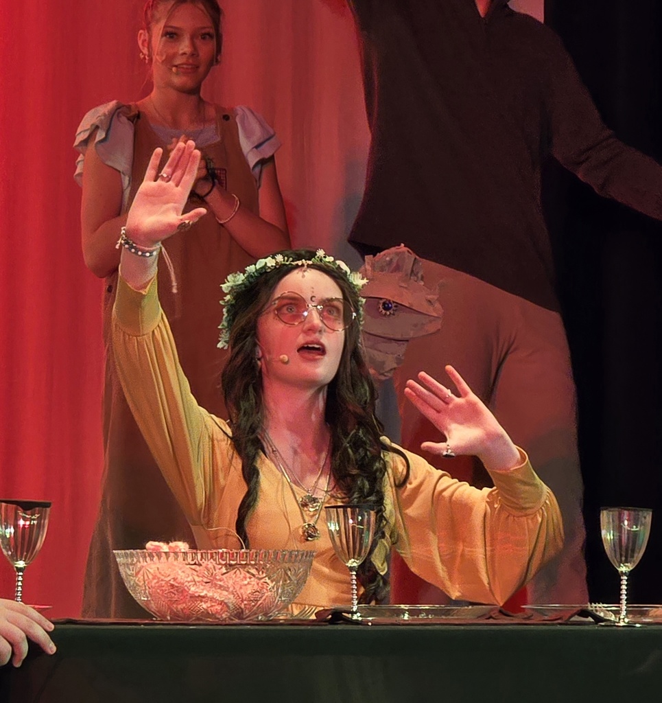 A woman in yellow waves at the audience; she has a flower crown and a microphone.