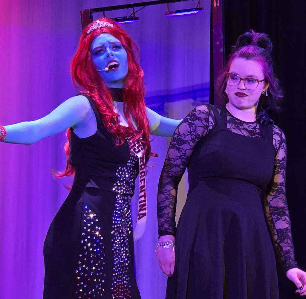 Two women, one with red hair and blue face paint, perform on stage. Both wear black dresses.