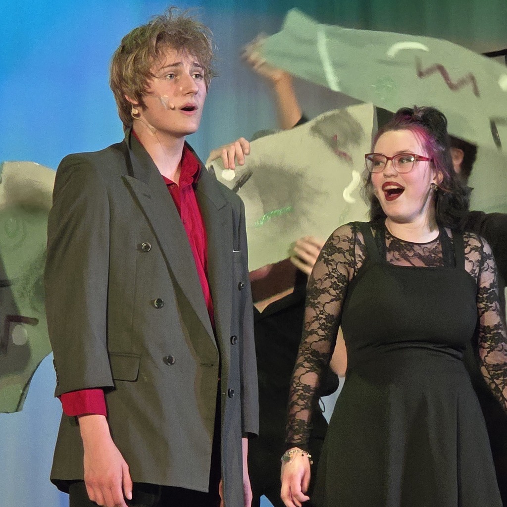 Two people on a stage; one in a gray jacket and red shirt, the other in a black dress with lace sleeves.