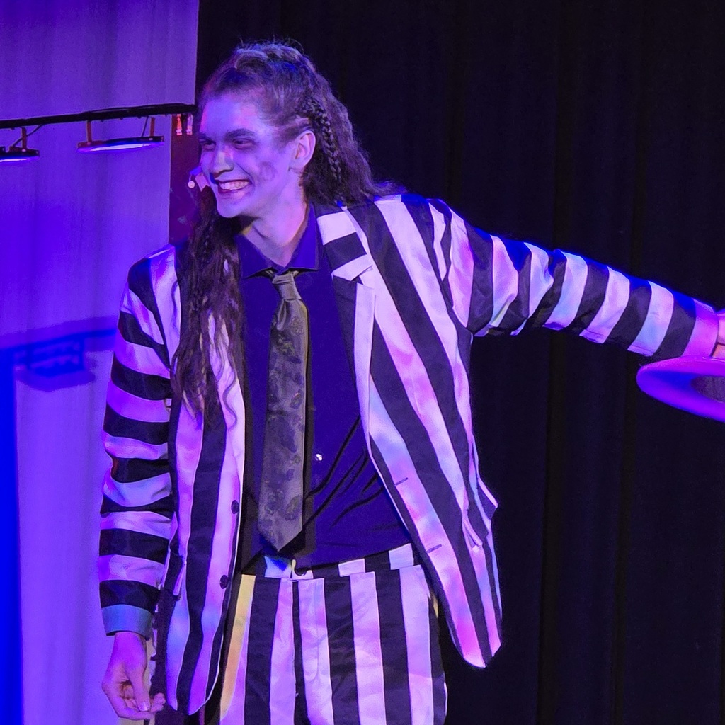 A person with long hair in a striped costume on a stage, smiling, and holding a prop.