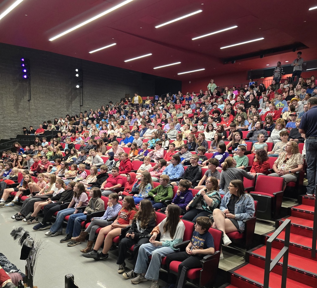 An indoor auditorium with many seated in red chairs, red steps, and a red ceiling with bright lights.