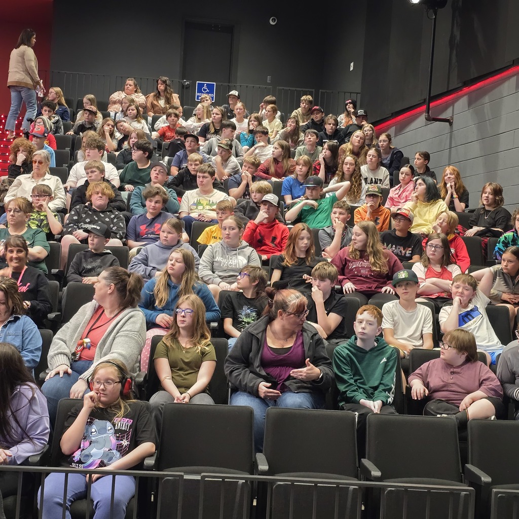 A large crowd of people seated in rows of black chairs in a theater with a red carpet.