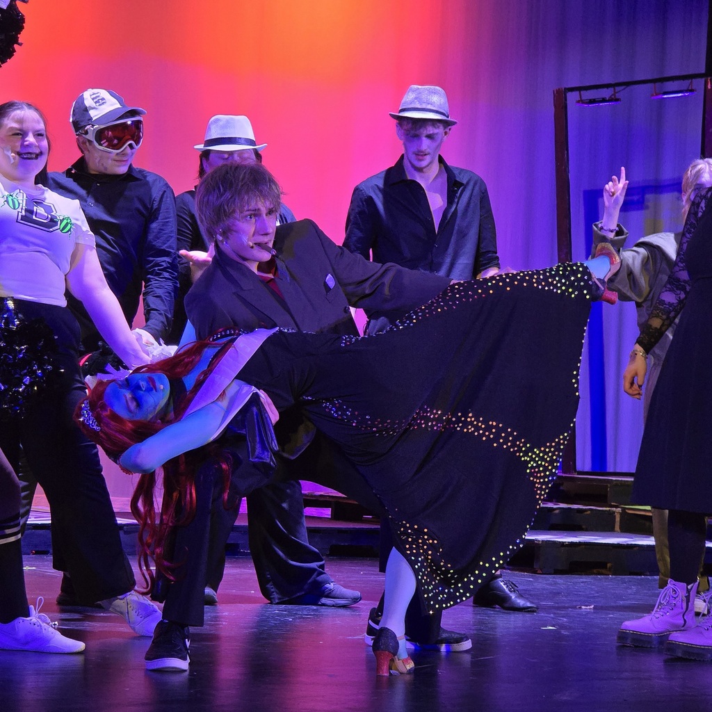 Dancers perform on a stage. A person in red makeup and a black dress spins while another person holds them.