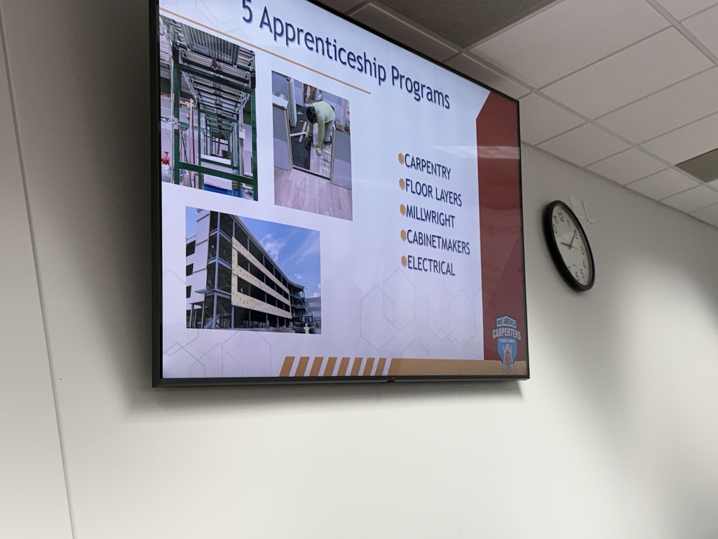 A large screen displays five apprenticeship programs with images of a building and a person working. A clock is on the wall next to the screen.