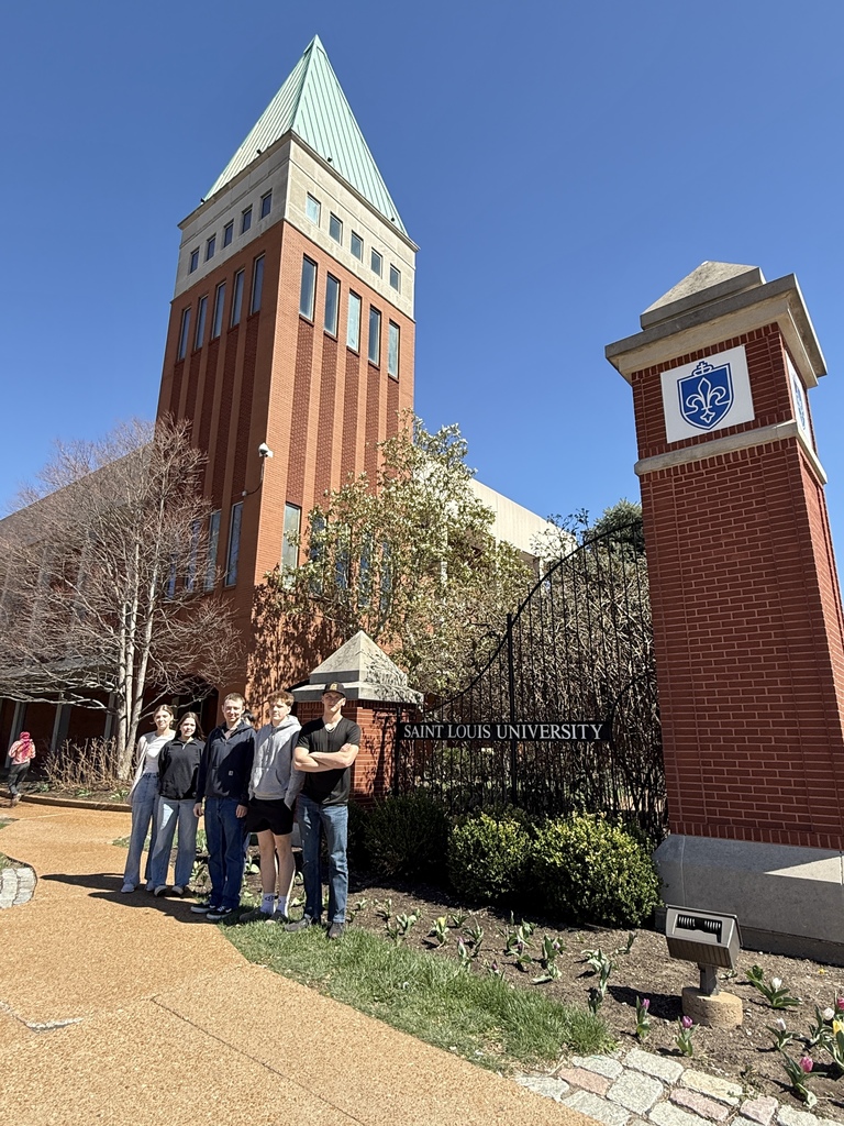 Five people stand in front of a building with a tower, brick walls, and a logo on the entrance.