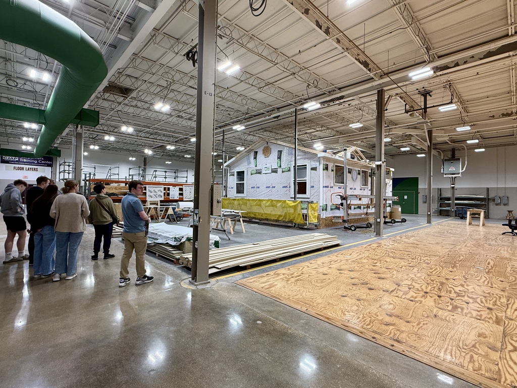 Several people stand around in a warehouse; a small house model is in the center. The floor has large wood sheets.