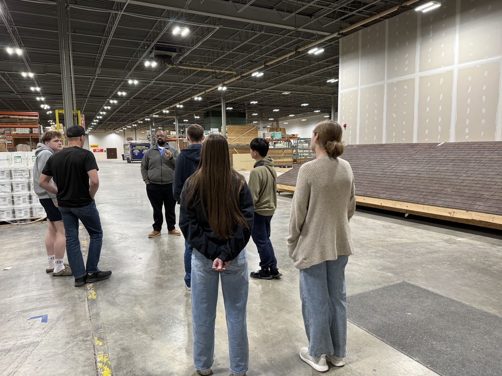 People in a warehouse with a floor of concrete, large unfinished wall, and stacked boxes.