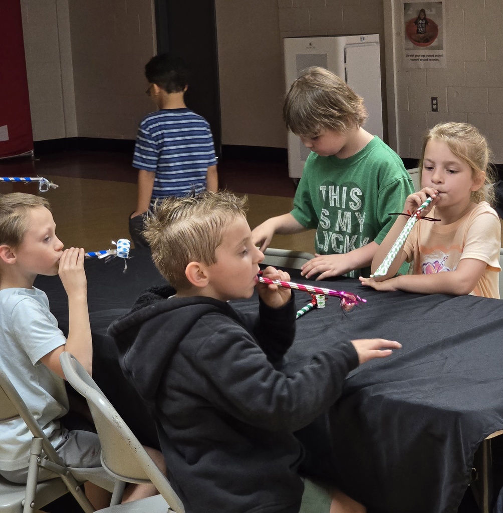 Five children sit at a table, blowing pink whistles. One wears a green shirt with a message, others wear casual clothing.