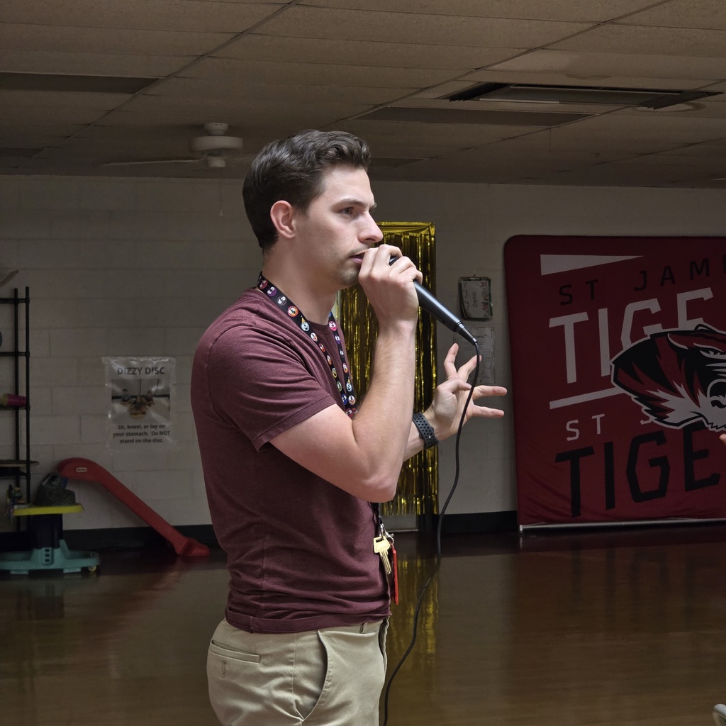 A man speaking into a microphone with a school logo in the background.