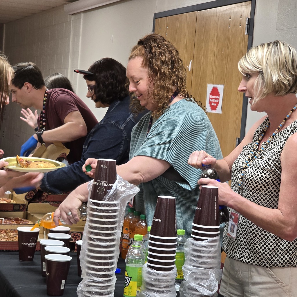 People in a room prepare and serve drinks at a table, including cups, bottles, and pizza.