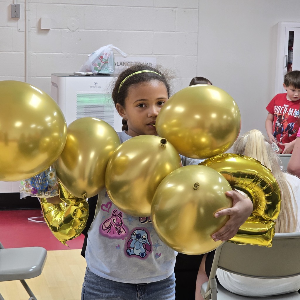A young girl holding a cluster of gold balloons stands in a room with chairs, a white wall, and a refrigerator.