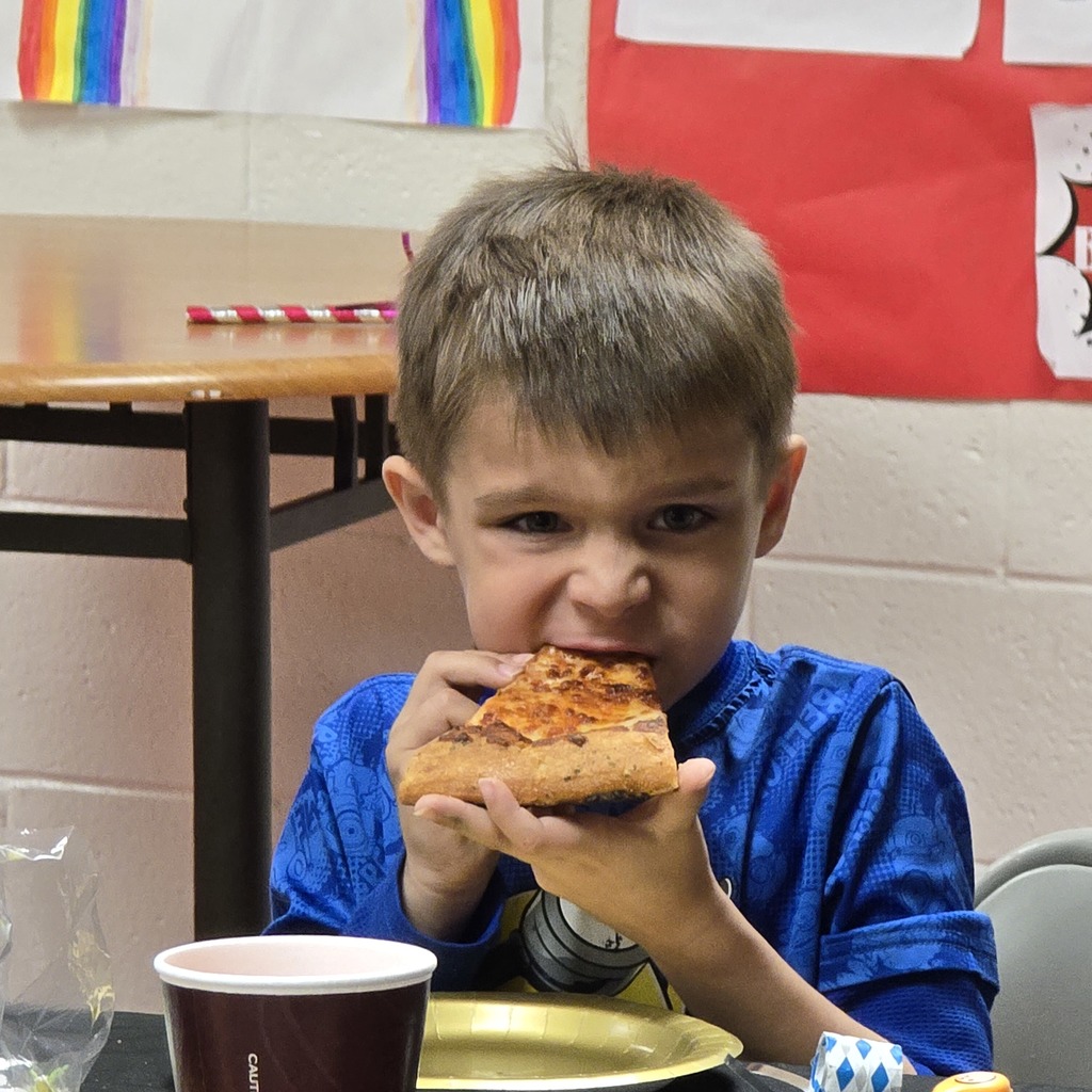 A boy in a blue shirt eats a slice of pizza, standing in front of a desk with a red banner.