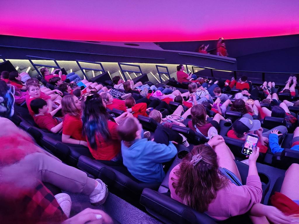 A theater with a large audience seated on chairs, some looking at their phones, with a purple ceiling.
