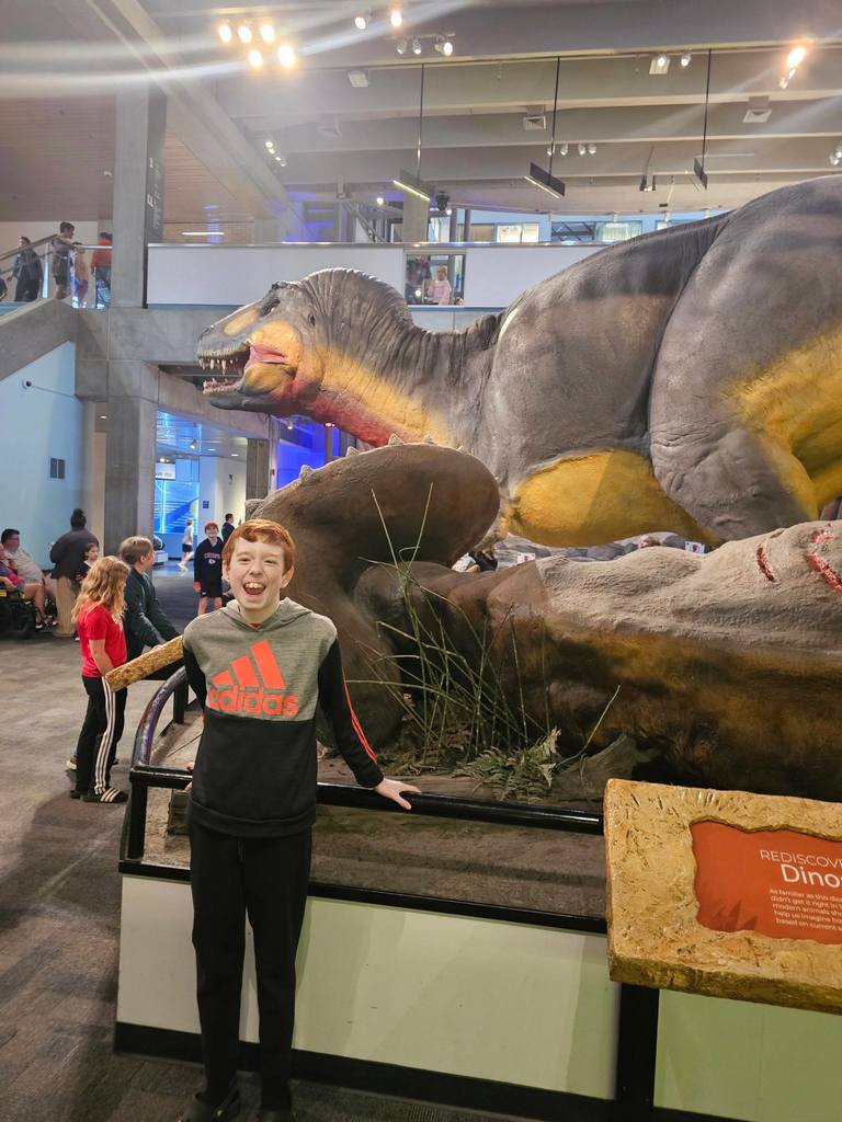 A boy stands in front of a large dinosaur statue inside a museum, with other visitors in the background.