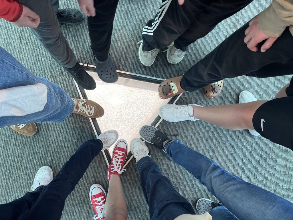 Group of people in sneakers and jeans standing in a circle around a square on the floor.