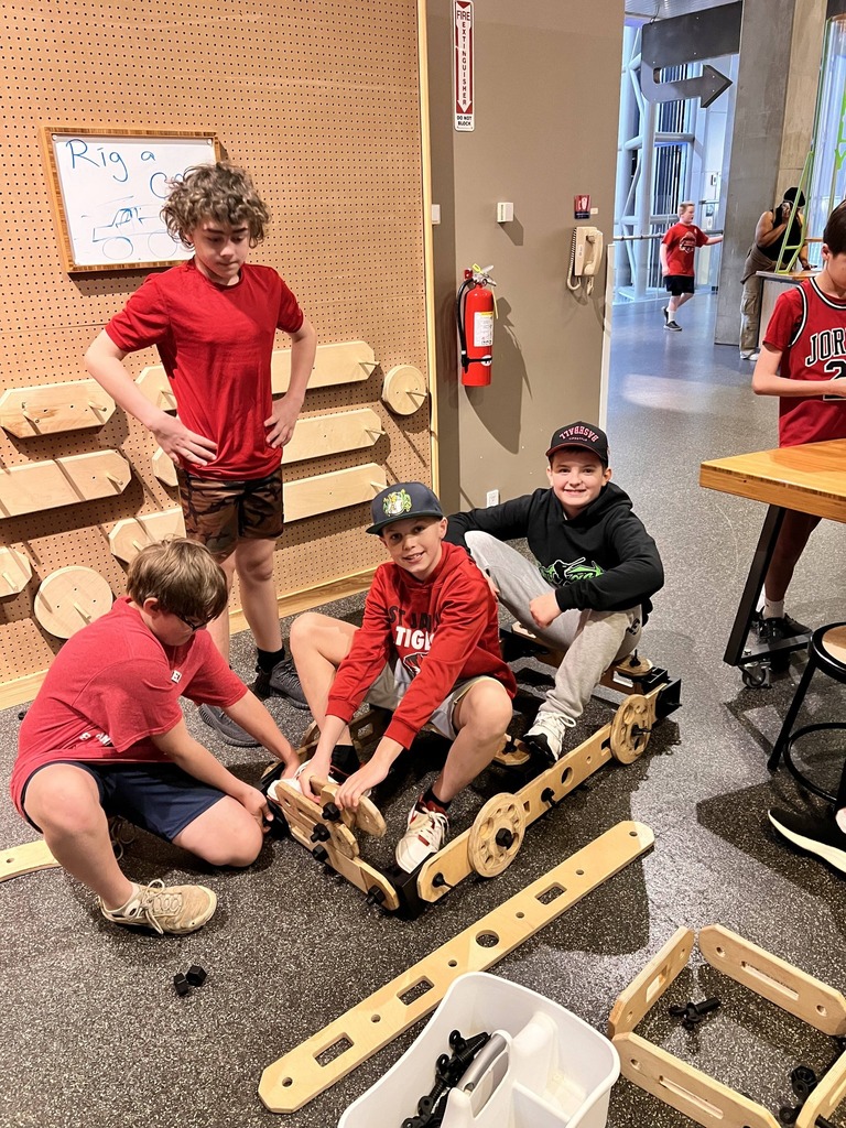 Children in red shirts are working on a wooden train project in a room with a wood wall.