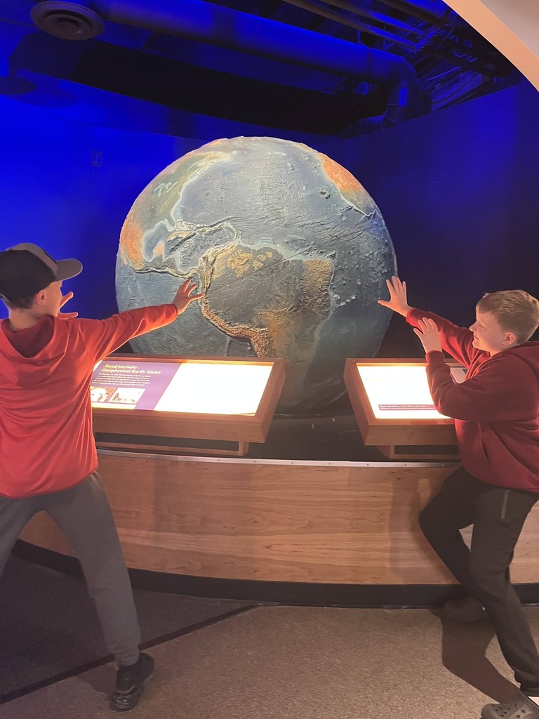 Two children engage with a large globe, pointing at regions on a lighted display. A blue wall and ceiling provide a science exhibit atmosphere.