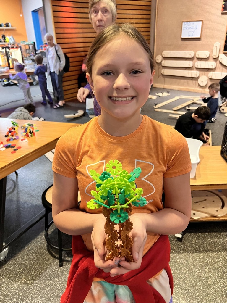 A girl in an orange shirt holds a tree made of colorful rubber bands. Behind her, there are tables with toys and people in a workshop.